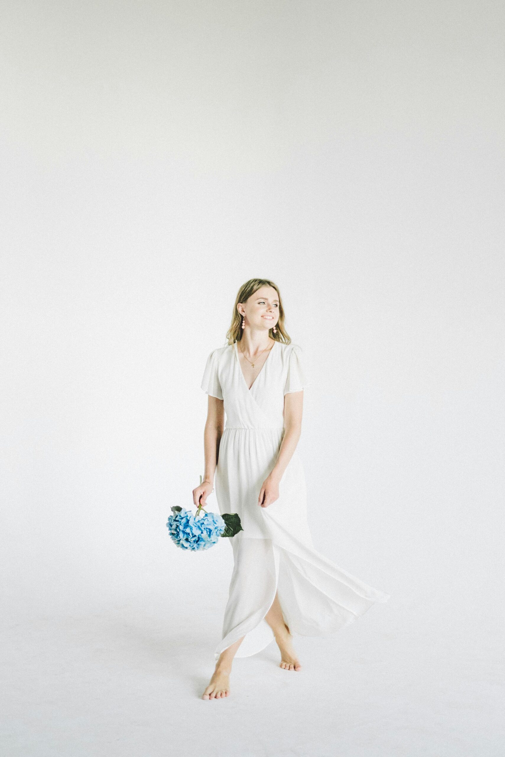 Elegant woman in white dress holding a blue hydrangea bouquet against a minimalist background.